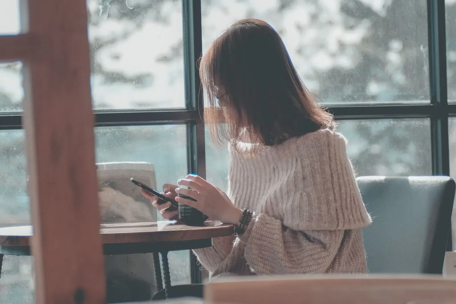 A woman sitting alone by a sunlit cafe window, holding a coffee and looking at her phone