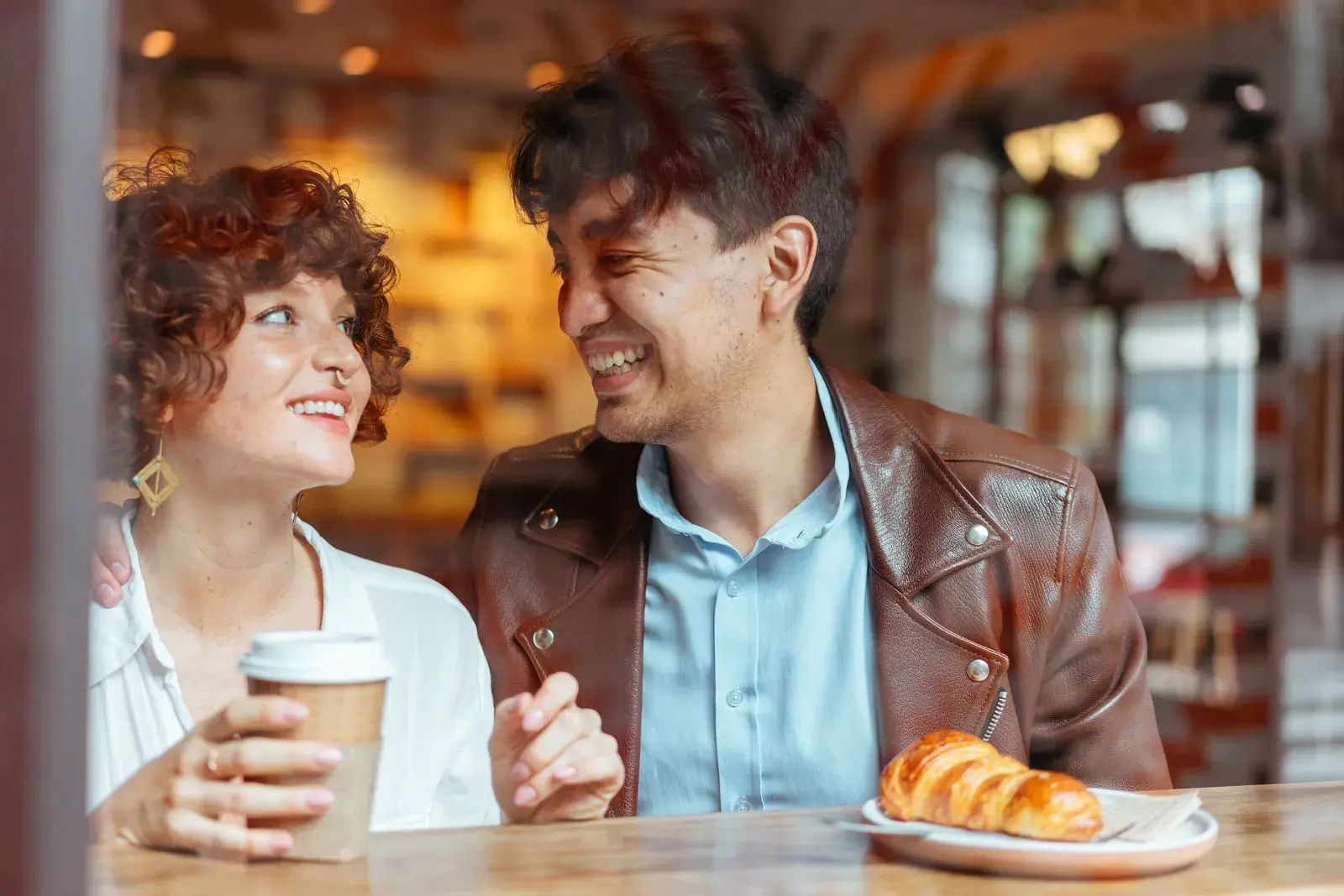 Two people laughing over coffee and a croissant at a sunny cafe